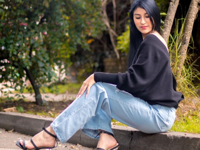 EmaHenderson está sentada em uma borda de concreto, vestindo um suéter preto, jeans azuis e saltos pretos, com cabelos pretos longos, batom vermelho e um leve sorriso, exibindo suas nádegas..