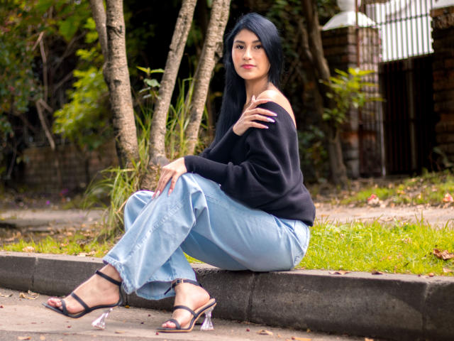 EmaHenderson with black off-shoulder top, light blue pants, and clear heels, sitting on concrete, hand on shoulder, black hair, red lips, green background, trees, and grass.