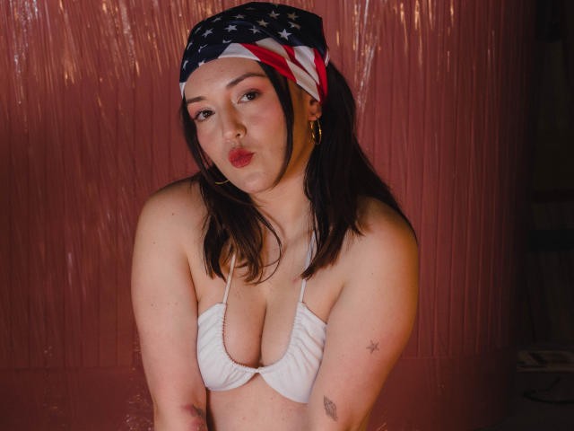 MollyMarmalade with medium tits in a white bikini top, wearing a red-white-blue bandana, pouting, has tattoos on her arms, and small hoop earrings, against a pinkish background.