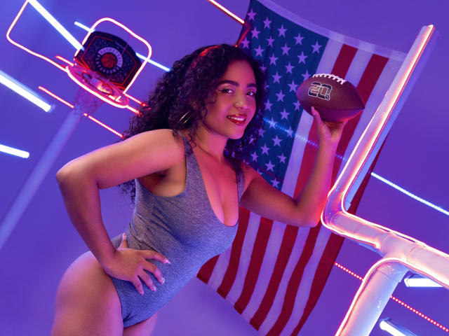 PaolitaKens with curly dark hair, wearing a grey one-piece, holding a football, smiling, neon lights, and American flag in background, showcasing her big ass and tits.