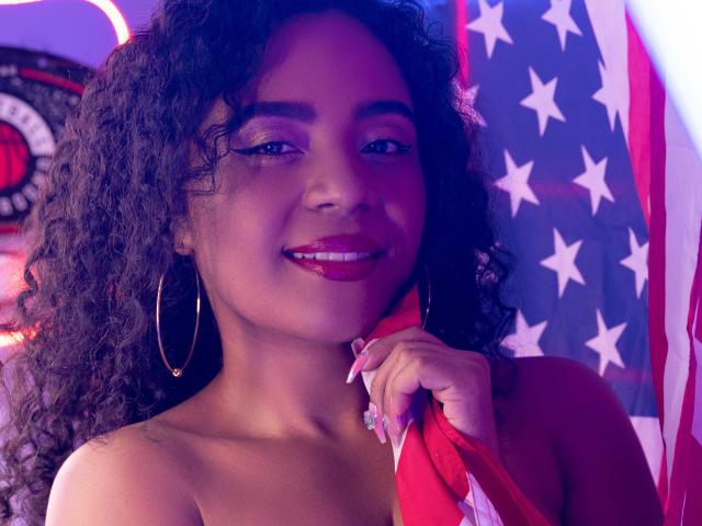 PaolitaKens with curly black hair, dark pink lipstick, and hoop earrings, holding a red flag, smiling, showing her tits, purple and pink lighting, American flag in background.