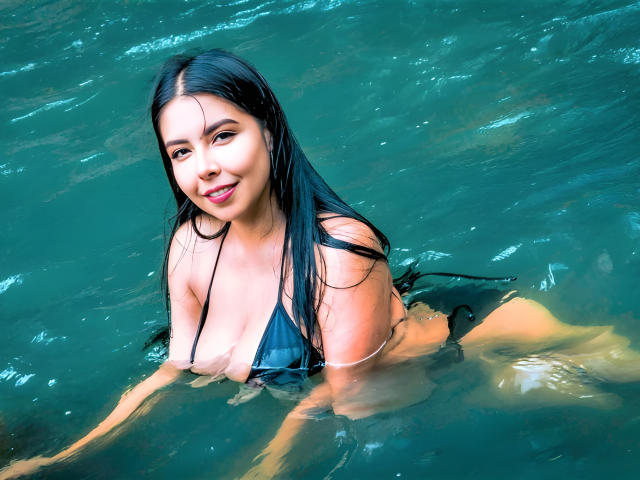 Samantha Klaus mit nassen, schwarzen Haaren, lächelnd in die Kamera, trägt einen schwarzen Bikini, der ihre großen, glänzenden Brüste zeigt. Sie steht am Ufer eines grünen Gewässers..