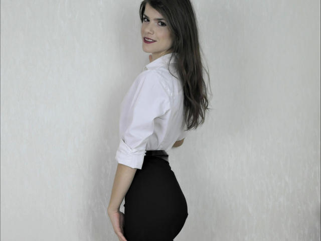 JustAlly-hot with long brown hair, white shirt, and black high-waisted pants, looking back with a smile, showing her butt. She's standing against a plain white wall.