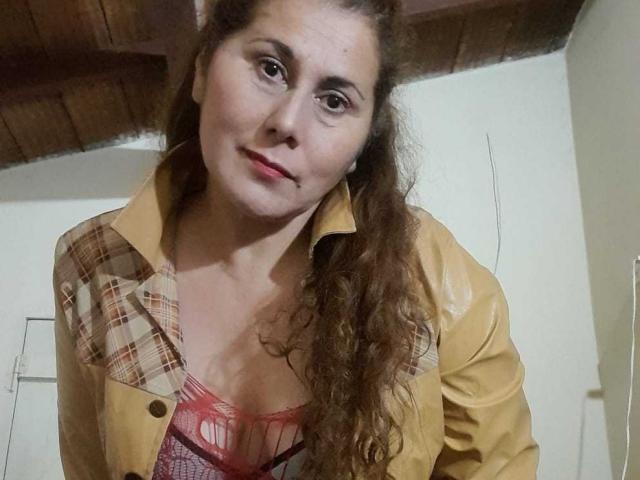 MirandaMature69 has curly brown hair, light skin, and wears a yellow jacket over a red lace bra, showing small tits and a slight smile. Her eyes are brown.