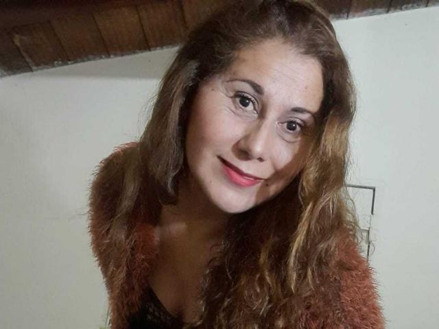 MirandaMature69 with wavy brown hair, red lipstick, and a fuzzy brown jacket, smiling at the camera. She's got a cute face and small tits, looking sexy.