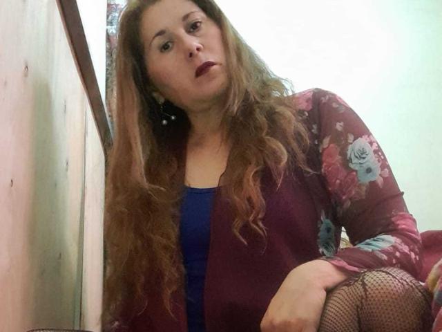 MirandaMature69 with wavy brown hair, purple floral robe, blue dress, and fishnet stockings, looking down with dark lipstick, sitting in a room, right hand on knee.