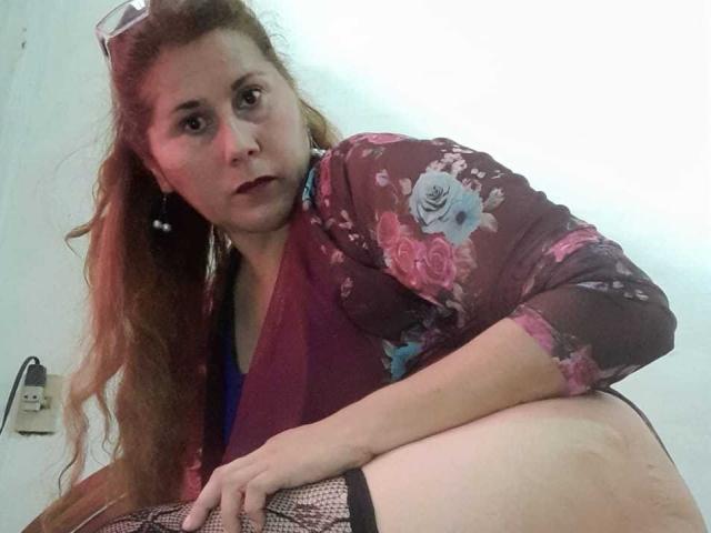 MirandaMature69 with long red hair, wearing a floral brown top, purple scarf, and black fishnet stockings, leaning forward with a serious expression, showing her bare ass.