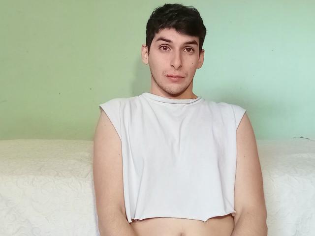 Alexxii with short dark hair, brown eyes, and light skin, wearing a white sleeveless crop top, showing a flat chest and toned abs. Simple green background.