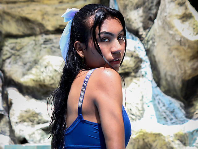 KattaleyaHorny with wet black hair in a blue swimsuit, showing her back and looking over her shoulder, her tits visible through the straps, nose ring, and blue bow.