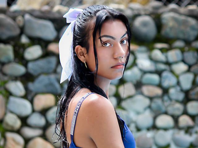 KattaleyaHorny with wet black hair, white bow, nose ring, and blue top, looking over shoulder, small tits visible, standing in front of a rock wall.