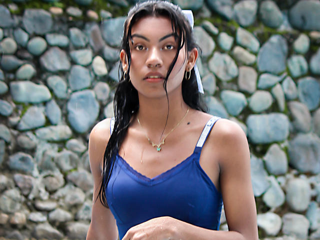 KattaleyaHorny has dark wet hair, a blue tank top showing small tits, white straps, gold necklace, and a serious face, standing in front of a stone wall.