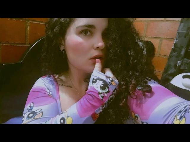 ReichelHott with curly black hair, pink and purple cartoon-patterned shirt, finger touching her slightly open mouth, looking up with a thoughtful expression, sitting against a brick wall.