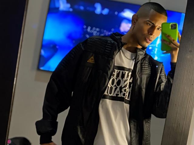 TybelMont with a short buzzcut, dark leather jacket, white tee, holding a green phone, standing in front of a blue TV, looking at a mirror reflection.