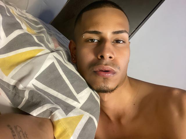 TybelMont with short hair, light brown skin, and a small facial piercing, lying shirtless, staring into the camera, holding a patterned pillow over his shoulder.