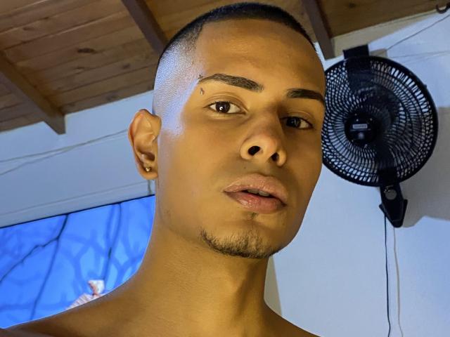 TybelMont, shirtless, has a clean-shaven face, short black hair, small facial scar above right eyebrow, and a small goatee. Black fan on white wall, wooden ceiling.