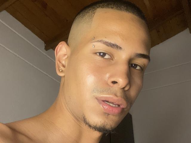 TybelMont's face, with short buzz cut, small nose piercing, ear stud, slight beard, glossy lips, and brown eyes, showing a neutral expression. Wooden ceiling above.