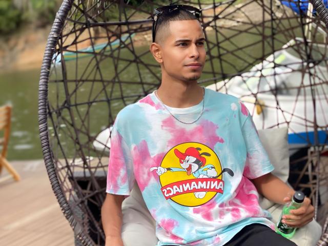 TybelMont with a cute, young face, short hair, and a light skin tone, wearing a pink and blue tie-dye shirt, sitting in a wicker hanging chair, holding a green bottle.