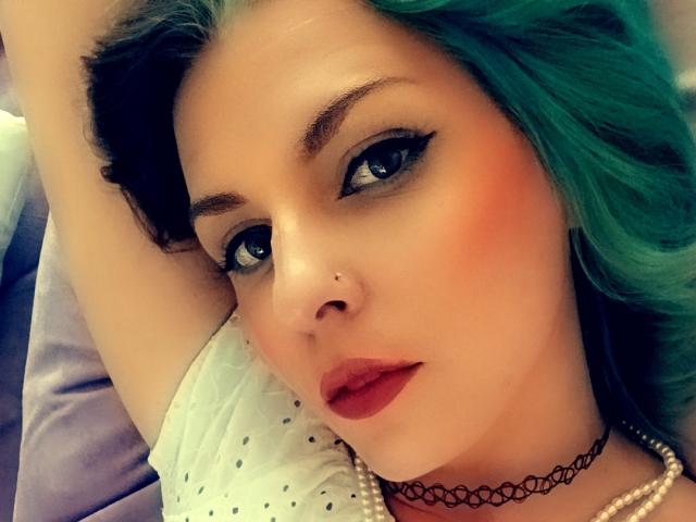 LunaLuxe with green hair, black eyeliner, red lipstick, and a nose piercing, lying down with arms above her head, wearing a black choker and white polka-dot shirt.