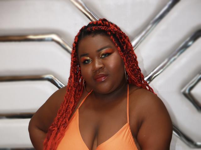 BritannyJhonson with red braids, orange bikini top, large boobs, shiny pink lips, green eyeshadow, and hoop earrings, looking down with a slight smile.