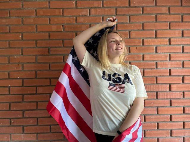 SallyVerda with blonde hair, holding an American flag, wearing a USA t-shirt, standing against a brick wall, smiling, right arm raised, flag draped over her left shoulder.