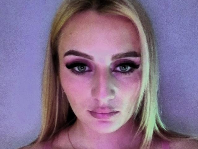 HesterBetta has blonde hair, pink eyeshadow, thick black eyeliner, green eyes, and pink lipstick. She's looking at the camera with a slight pout. Her tits are not visible.