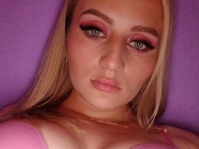 HesterBetta with pink makeup, blonde hair, green eyes, pink lipstick, and pink bra, looking at the camera with a neutral expression. Her tits are slightly visible.