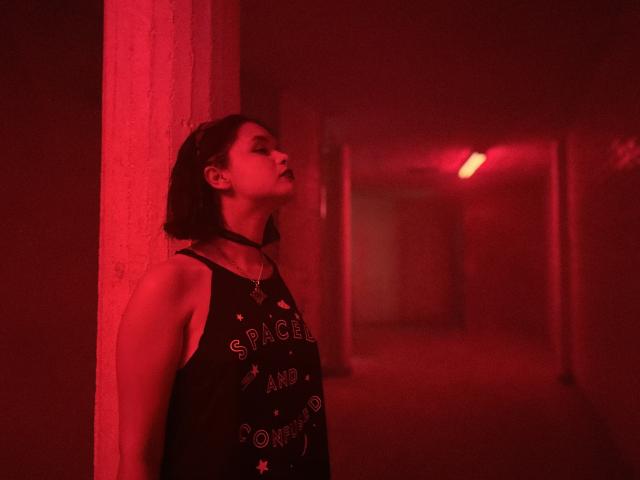 CatarsisNous with short black hair, red-lit corridor, leaning against wall, black tank top, choker, looking up, red light, red shadows, slim build, slight muscle definition.