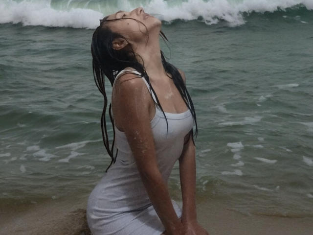 KiriamGomez, com o cabelo preto molhado, a cabeça inclinada para trás e os olhos fechados, vestindo um vestido branco, molhado e justo ao corpo, sentada em uma praia com ondas do oceano ao fundo. Seus seios estão visíveis..