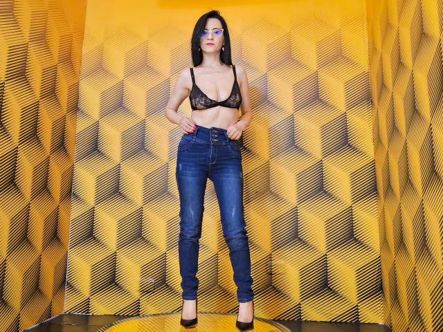 AlanaMoneth with black lace bra, showing her tits, wearing blue jeans and black heels, standing with hands on hips, against yellow, patterned wall.