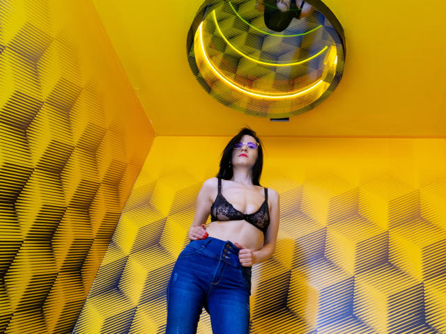 AlanaMoneth com um sutiã de renda preta que mostra seus seios, vestindo jeans azuis, em pé em um quarto amarelo com padrão geométrico, olhando para cima para o luminária circular amarela no teto..