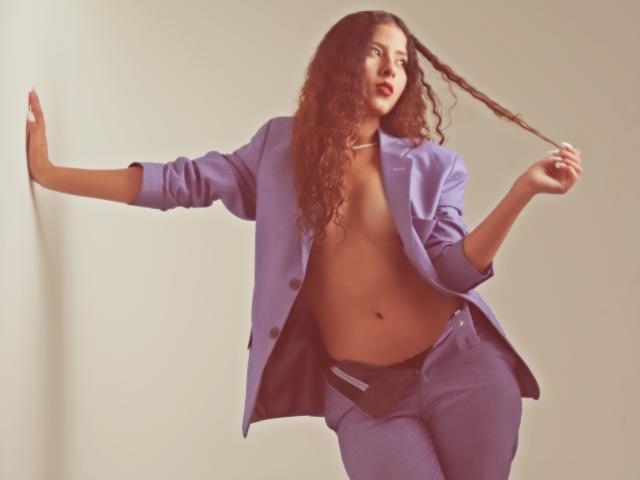 Elena Santini, com cabelos castanhos cacheados, vestindo um blazer roxo e com os botões abertos, expondo seus seios nus e roupa íntima preta, segurando o cabelo com a mão direita..