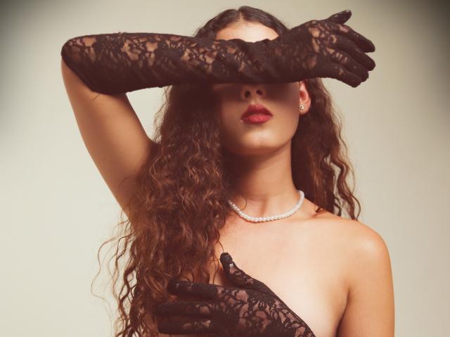 Elena Santini mit welligem, braunem Haar, trägt schwarze Spitzenhandschuhe, bedeckt ihre Brüste mit einer Hand, hat roten Lippenstift, eine Perlenkette und trägt kein Oberteil, während sie ihren Arm hebt..