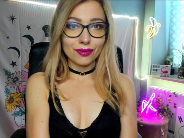 Blonde HeartHeidi, avec des lunettes, du rouge à lèvres rose, un collier noir et un haut noir décolleté, montrant ses seins. Des lumières néon et des fleurs sont visibles en arrière-plan. Elle sourit à l'appareil photo..