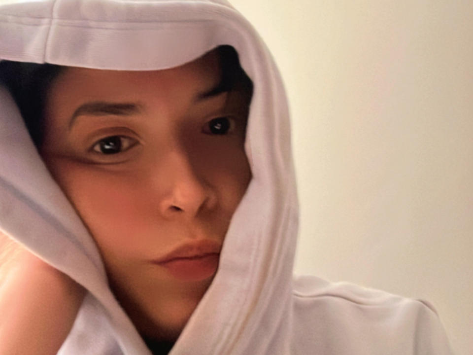 Melix with a serious look, wearing a white hoodie, hood up, exposing her face and brown eyes, has her right hand near her head, in a soft-lit selfie.