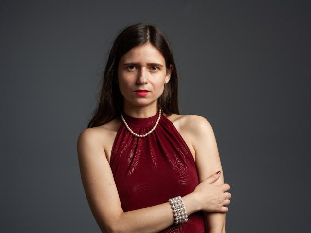 MartaLeee with long brown hair, red halter dress, pearl necklace, and bracelet, arms crossed, looking serious, gray background, tits partially visible, red lipstick, dark eyes, slight frown.