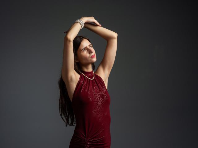 MartaLeee hat die Arme erhoben, trägt ein rotes Kleid mit einem gemusterten Brustbereich und ein Armband am Handgelenk, hat langes, braunes Haar und rote Lippenstift..