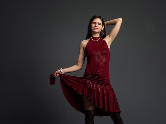 MartaLeee in a red dress with a ruffled skirt, holding it out, showing her thighs. She's wearing black stockings and a bracelet, with one arm raised.