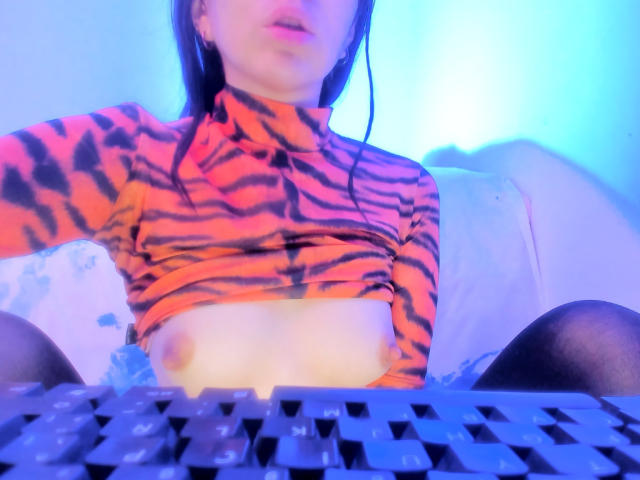 FairyBeauti with taut tiger-striped top, exposing small bare tits, sitting with legs apart, blue light, black keyboard in foreground, mouth slightly open, long dark hair.