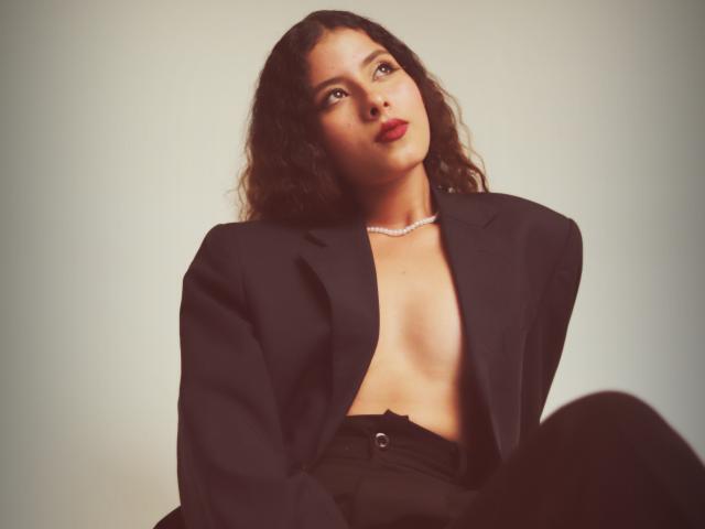 ElenaSantini with curly dark hair, wearing an open black jacket, exposing her tits, black pants, and a diamond necklace, looking up with red lipstick.