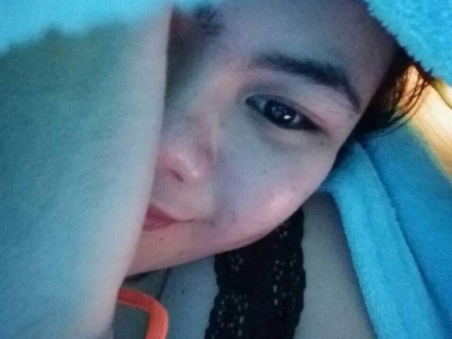 ChinitaPrincess's face with blue towel covering her head, dark eyebrows, black lace top, and small nose, looking slightly to the side with a subtle smile, arms partially visible.