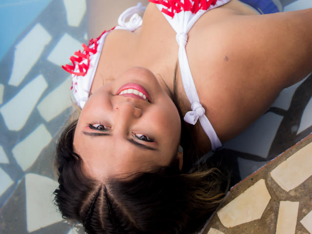 GraceJansen with dark brown hair, braided, smiling upside down, wearing a red and white polka dot bikini top, showing her tits, in a pool, with blue water.