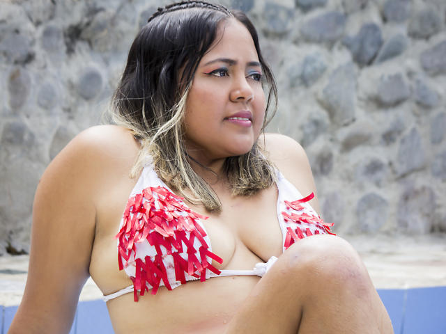 GraceJansen with medium brown skin, black and blonde ombre hair, wearing a red and white fringed bikini, showing her big tits, sitting with a slightly annoyed expression.