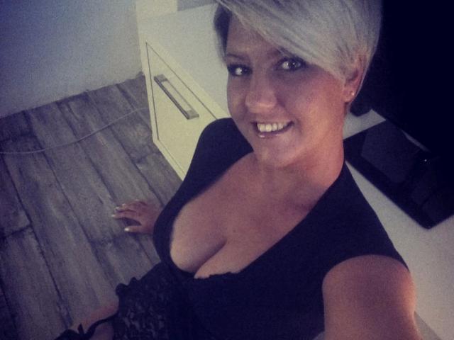 NatiLove-hot has short blonde hair, smiling, wearing a low-cut black top showing her big tits, standing with a wooden floor, and a white cabinet in the background.
