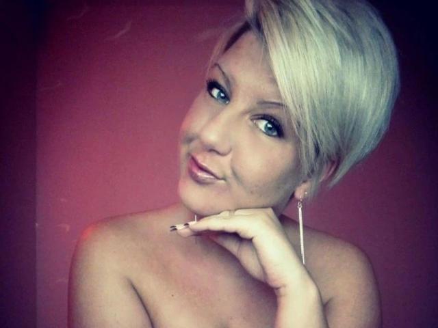 NatiLove-hot has short blonde hair, blue eyes, and is smiling with her hand on her chin. She's topless, showing her bare shoulders and a small, cute smile.