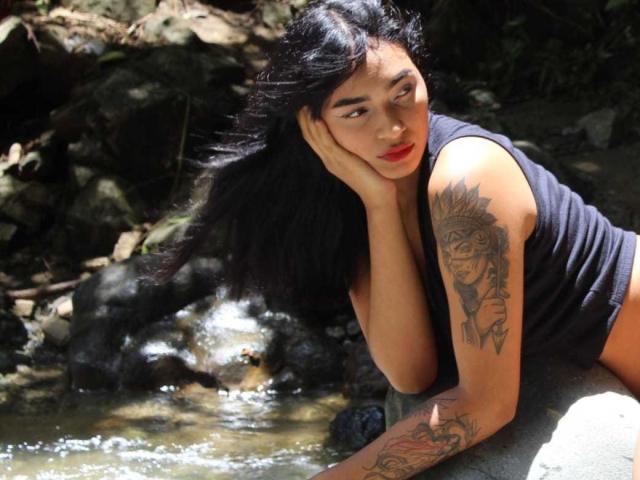 MelodyAsian with long black hair, red lipstick, black sleeveless top, and tattoos on her right arm, leaning by a stream, hand on cheek, looking down, sunlight on her face.