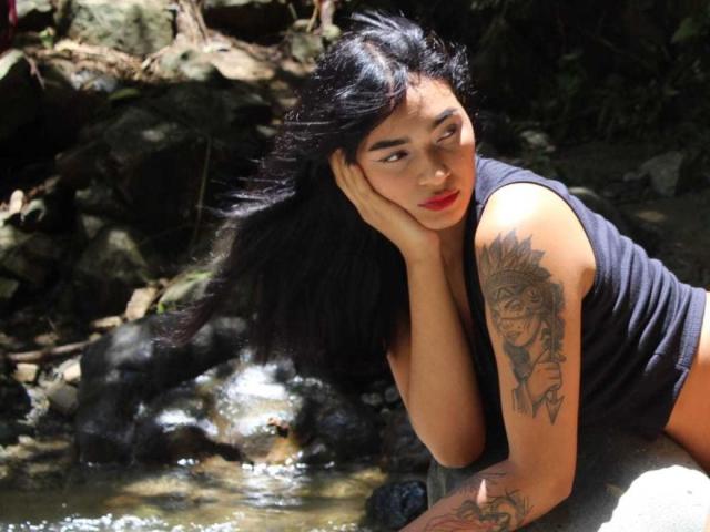 MelodyAsian with long black hair, red lipstick, black tank top, tattoo on right arm, hand on cheek, sitting by rocky stream, looking off to the side, sunlight.