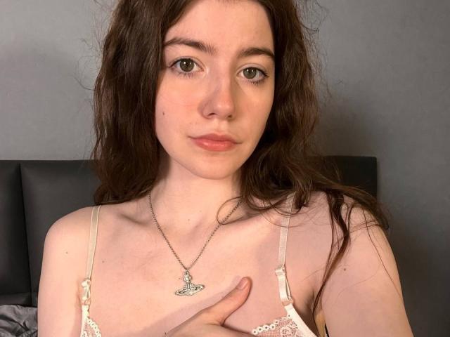 VinnaSkyHott with pale skin, brown wavy hair, and green eyes, wearing a white lace bra, silver butterfly necklace, and touching her chest, showing small tits.