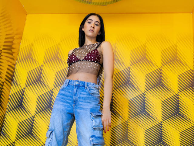 NicoColeman with dark hair, wearing a sheer red polka-dot bralette, showing small tits, and blue ripped jeans, standing against a yellow geometric-patterned wall.