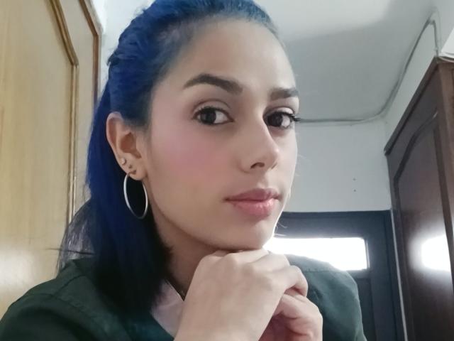 AnaisHilton has blue hair, hoop earrings, and pink blush, resting her chin on her hand with a subtle smile. Her tits are not visible. She's in a wooden room.