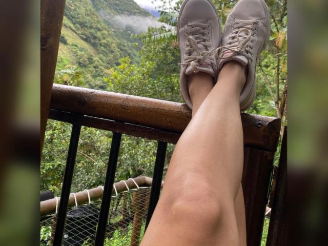 Kendra Milers Beine, mit heller Haut, trägt graue Sneakers, die an den Knöcheln gekreuzt sind, und ruhen auf einem hölzernen Geländer, umgeben von einem üppigen, grünen Wald..